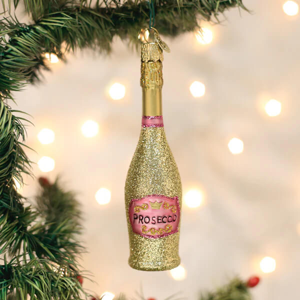 A glittery gold prosecco bottle ornament with pink label and foil cap hanging from a christmas glass ornament tree branch