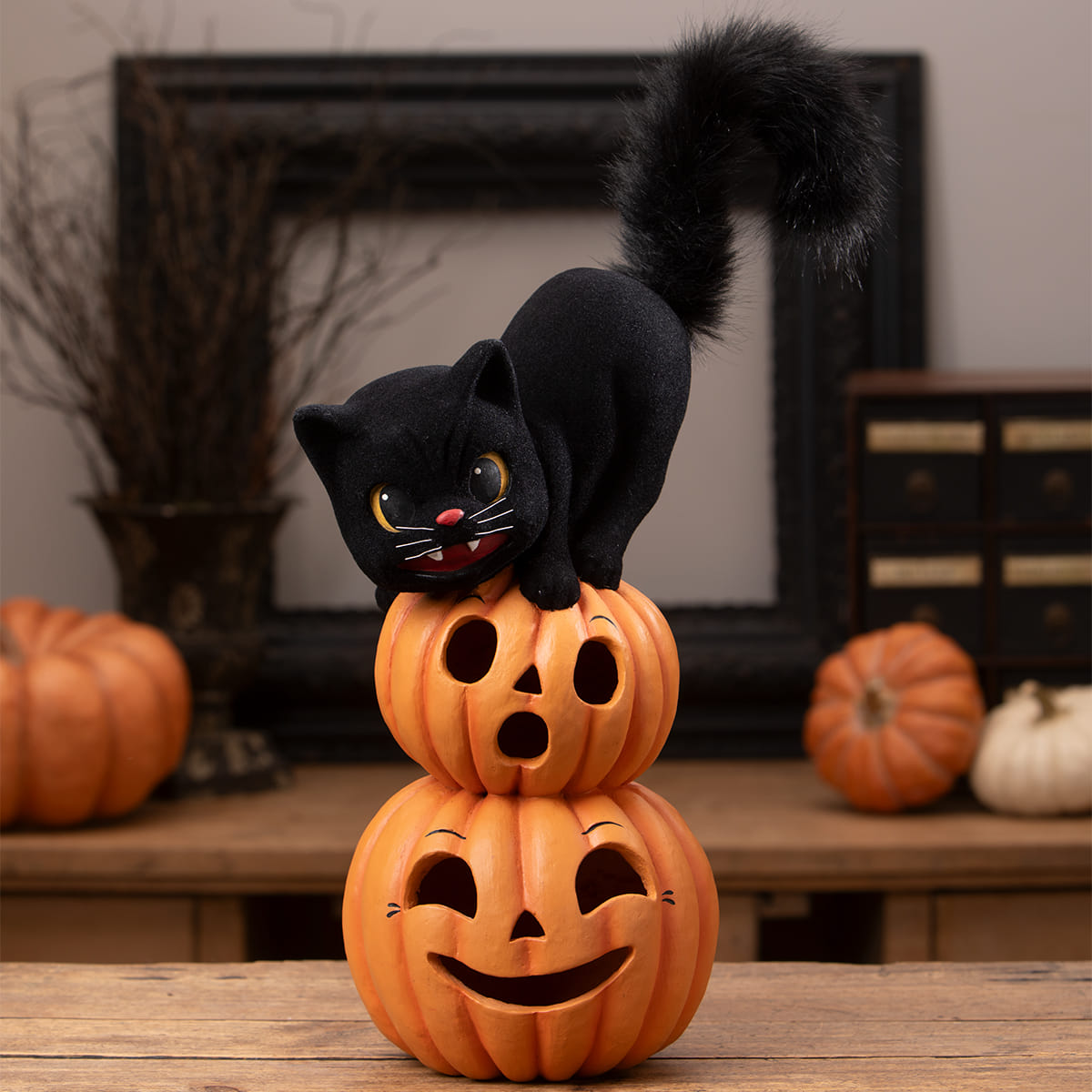 A black plush halloween cat with a fluffy tail perched on two stacked orange halloween pumpkins and gourds figurines