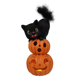 Black plush cat with fluffy tail sitting on stacked orange halloween pumpkins with smiling and surprised faces for halloween cats and gourds decor