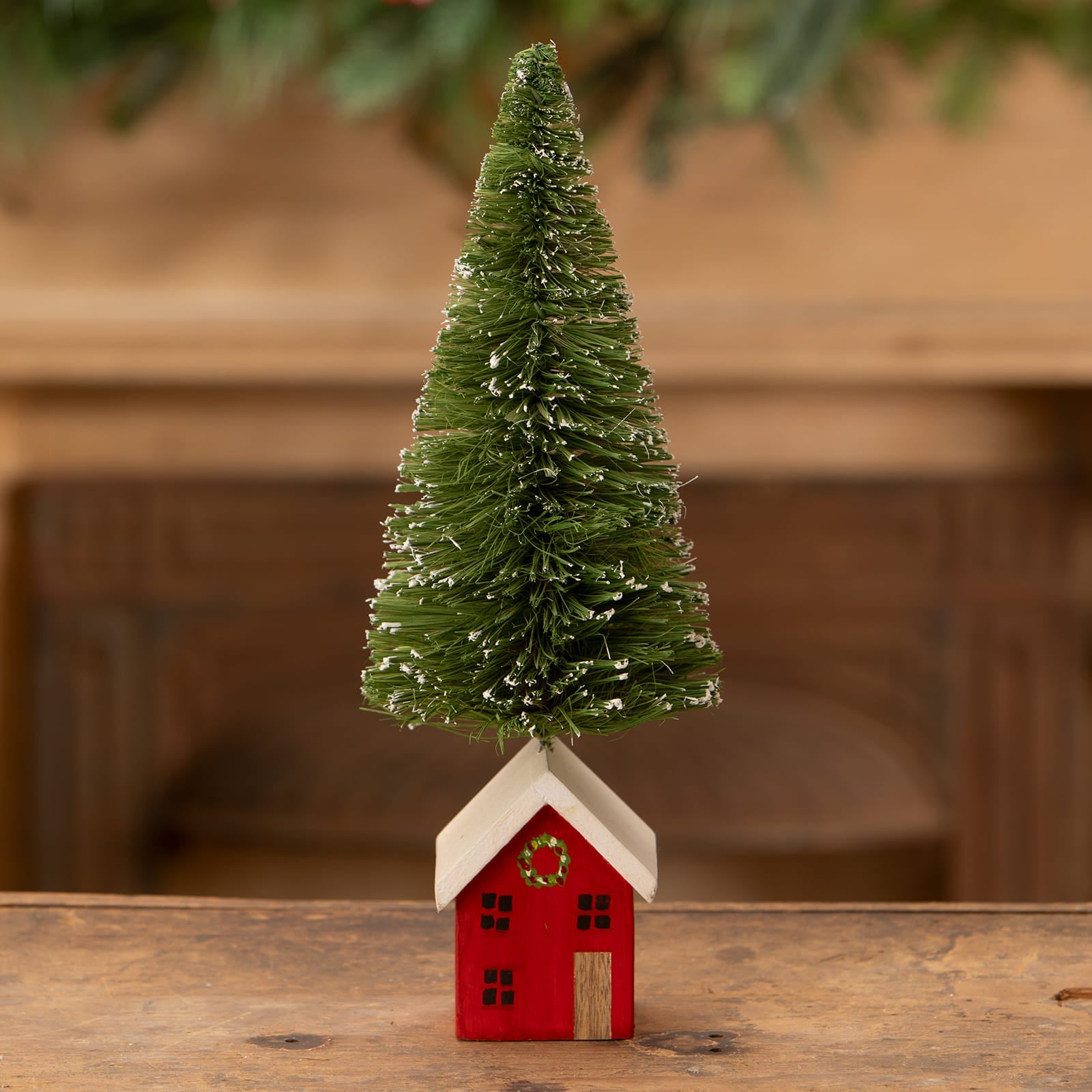 A green bottle brush tree with white tips sits on a red and white wooden house ornament in bethany lowe christmas trees