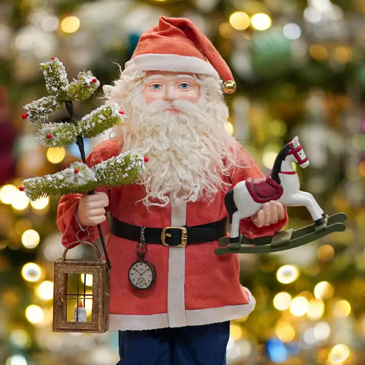 Decorative Santa Claus figure holding a tree, lantern, and rocking horse against a blurred Christmas tree background.