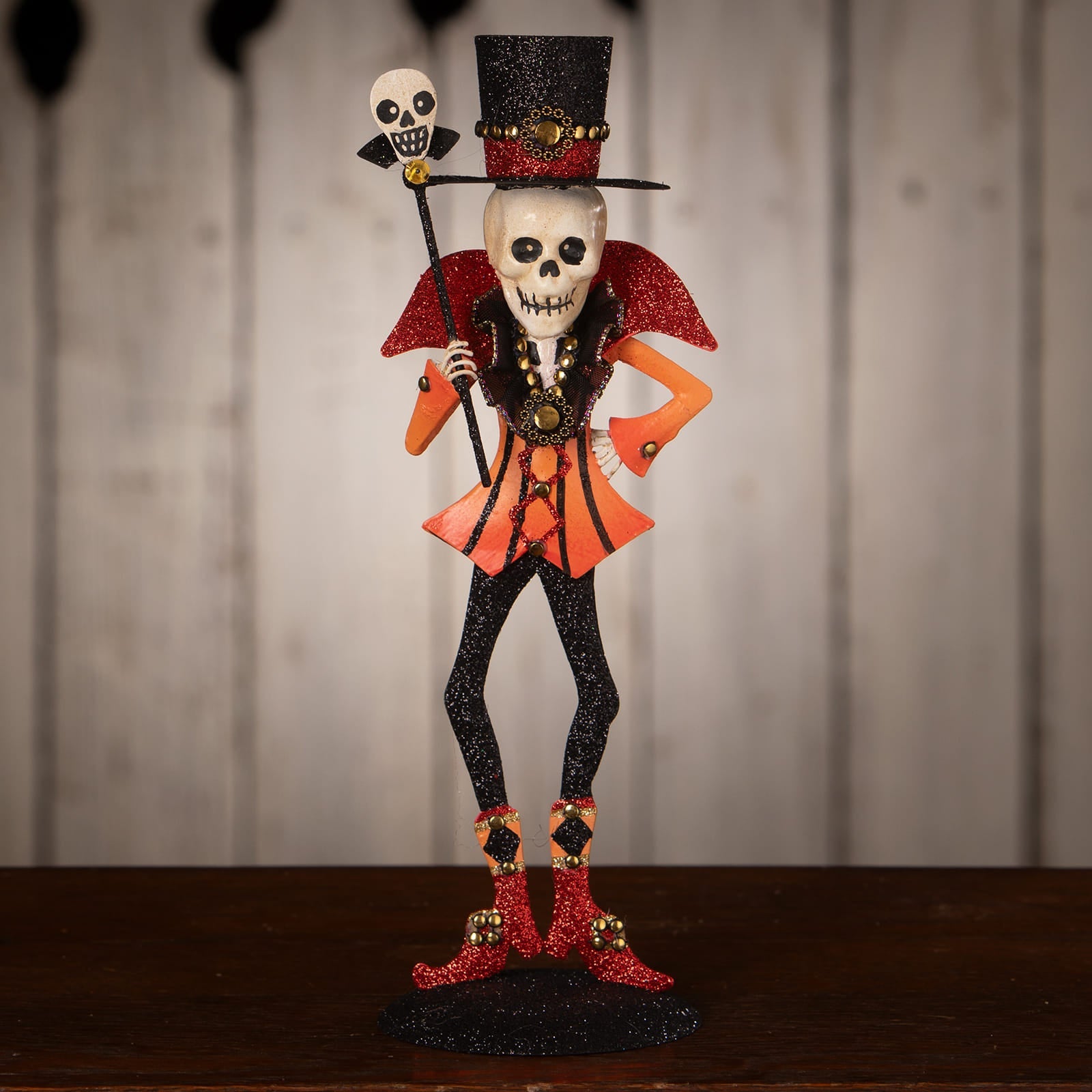 A glittery skeleton decor figure dressed as a ringmaster in a black top hat, orange cape, and red boots with gold studs