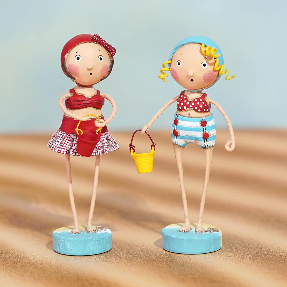 Two whimsical lori mitchell sandy beach figurines in red polka dot swimsuit and blue striped shorts on blue bases