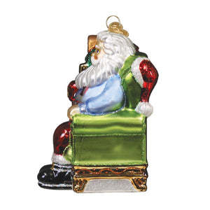 Glass santa ornament with white beard red and black suit blue shirt and gold glittery details on green chair