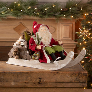 Santa on log sled figurine by bethany lowe designs in red and green holding snow-dusted pine branches on white sled