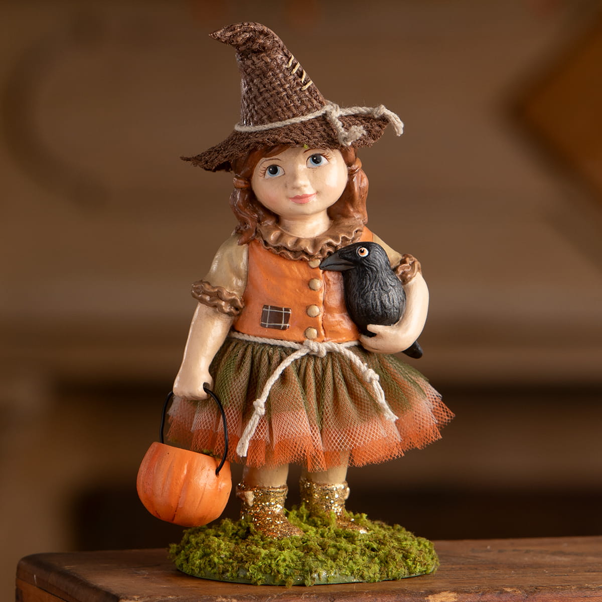 A charming scarecrow figurine of a girl in orange and brown holding a black crow and a pumpkin bucket for autumn décor
