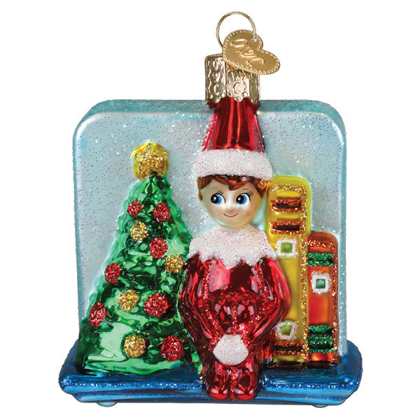 Red glass scout elf storytime ornament with white trim beside green glittery christmas tree and colorful books