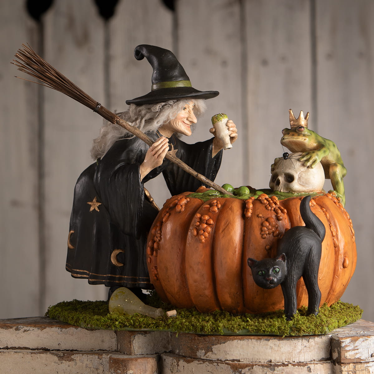 A halloween witch figurine in black robe with gold stars holding broomstick beside pumpkin with crowned frog skull prince potion