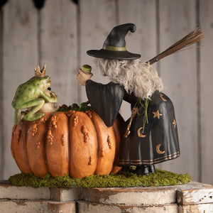 A whimsical halloween witches figurine of a black hat witch holding a green prince potion beside a pumpkin with a crowned frog