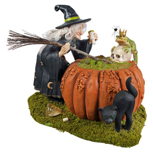 A whimsical halloween witch figurine in black with green trim holding a broomstick beside a pumpkin cauldron for prince potion