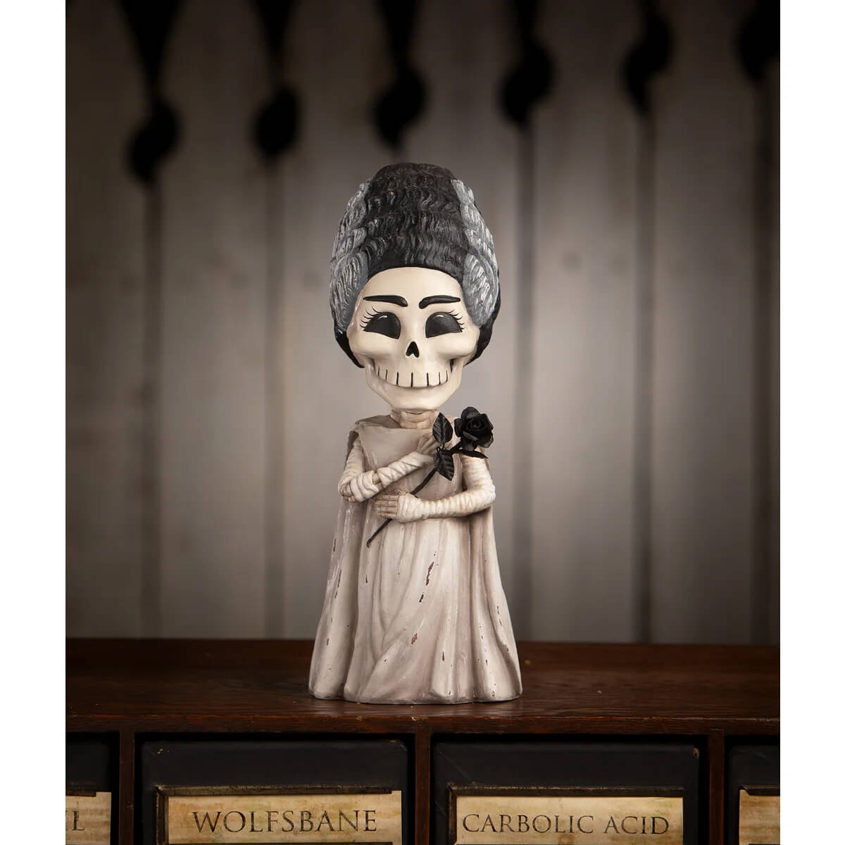 A shelley skelly figurine with a white skull face, black and gray hair, off-white gown, holding a black rose