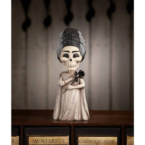 A shelley skelly figurine with a white skull face, black and gray hair, off-white gown, holding a black rose