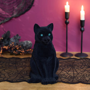 A sitting flocked green-eyed black cat plush figurine with glowing turquoise eyes on a lace table runner