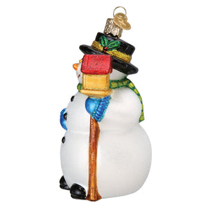 A festive glass snowman ornament with a black top hat adorned with green holly, wearing a blue scarf and holding a red-and-gold gift box on its shoulder.