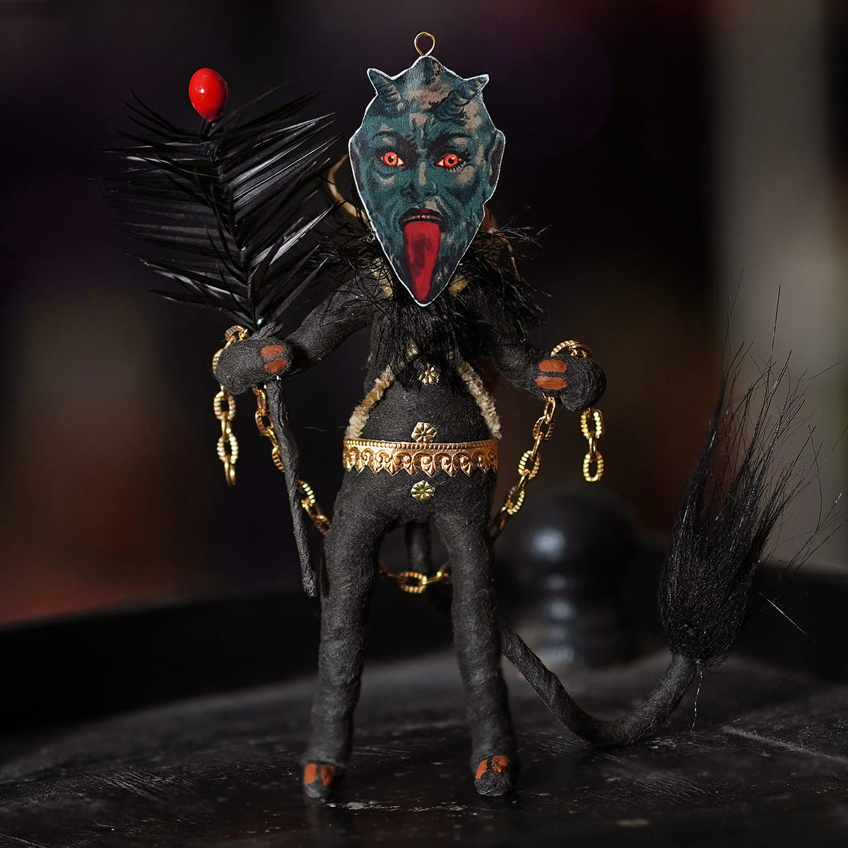 A spun cotton black krampus freestand ornament with a dark teal face, red eyes, black fabric, and gold chain details