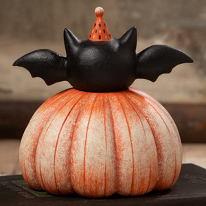 A black bat figurine with spread wings wearing a polka-dotted orange party hat on an orange pumpkin base for halloween pumpkins and gourds decor
