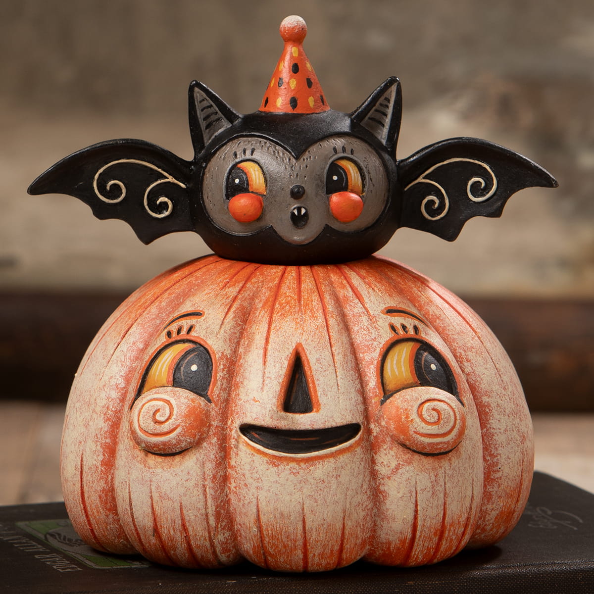 A halloween-themed ceramic figurine called squatty pumpkin batty with a smiling orange pumpkin and a black bat party hat perfect for halloween pumpkins and gourds decor