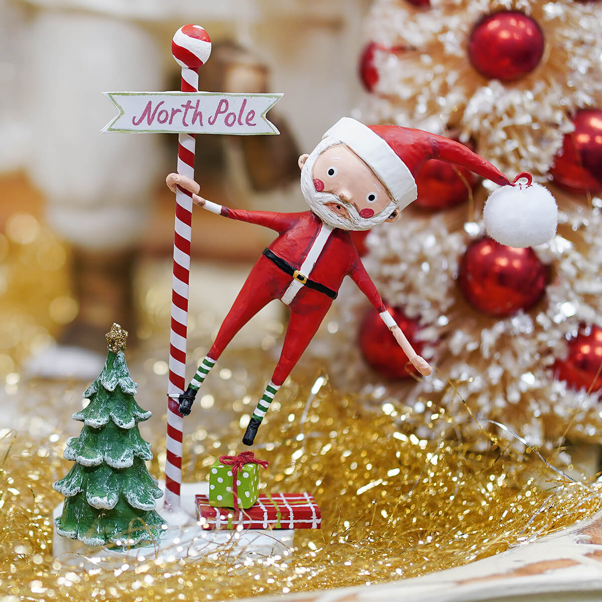 Red and white santa claus figurine holding north pole candy cane next to green tree and gifts christmas decorations ornaments