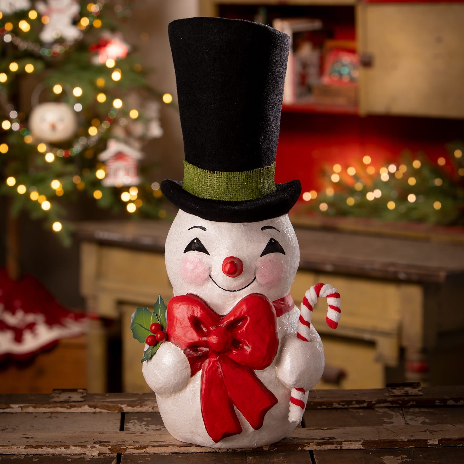 White ceramic snowman figurine with black top hat green band holding red bow and candy cane by bethany lowe designs