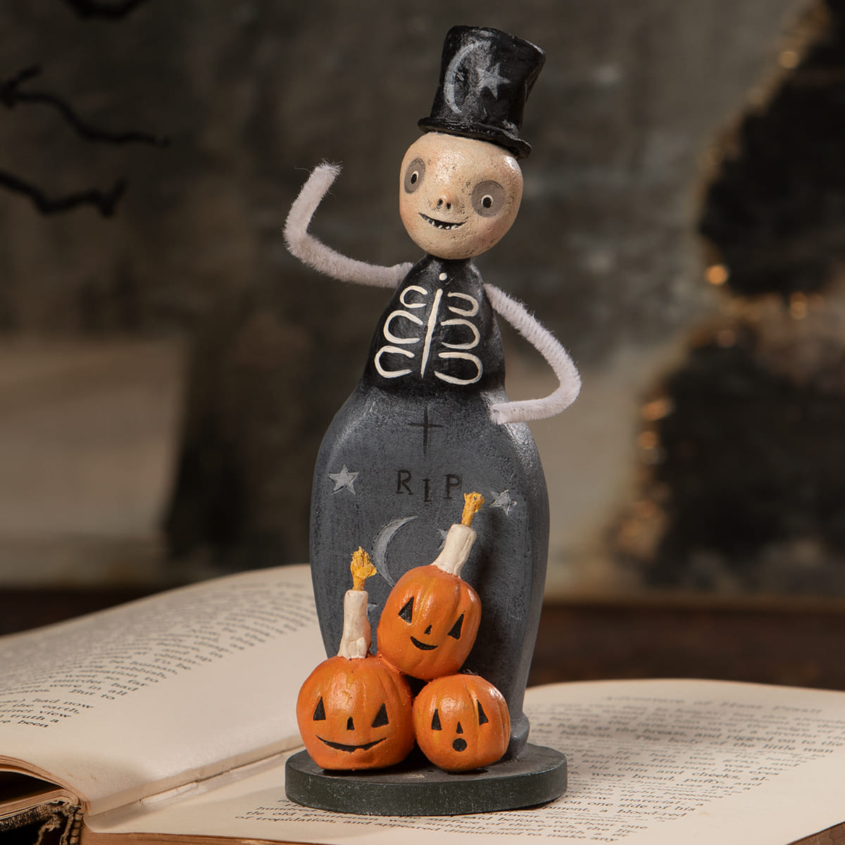 Tombstone trickster halloween figural decor with a whimsical skeleton in black vest and hat beside three jack o lanterns