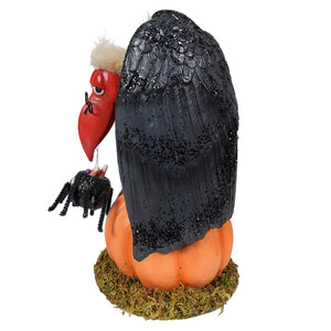 A black glittery vulture figurine with a red beak and white tufted head perched on an orange pumpkin base treats for the buzzard