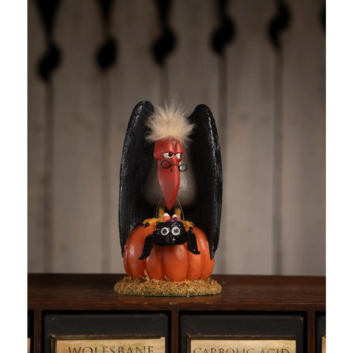 A whimsical black vulture figurine with white feathered crest and red beak perched on an orange pumpkin with a black spider in treats for the buzzard