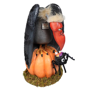 A black and orange halloween-themed vulture figurine with a red beak and fuzzy white hat perched on a pumpkin treats for the buzzard