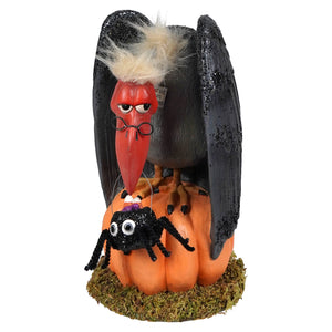 A black glittery vulture with a red beak and glasses perched on an orange pumpkin with a small spider for treats for the buzzard