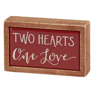 Wooden-framed decorative sign with red background displaying ’TWO HEARTS ONE LOVE’ text in white lettering.