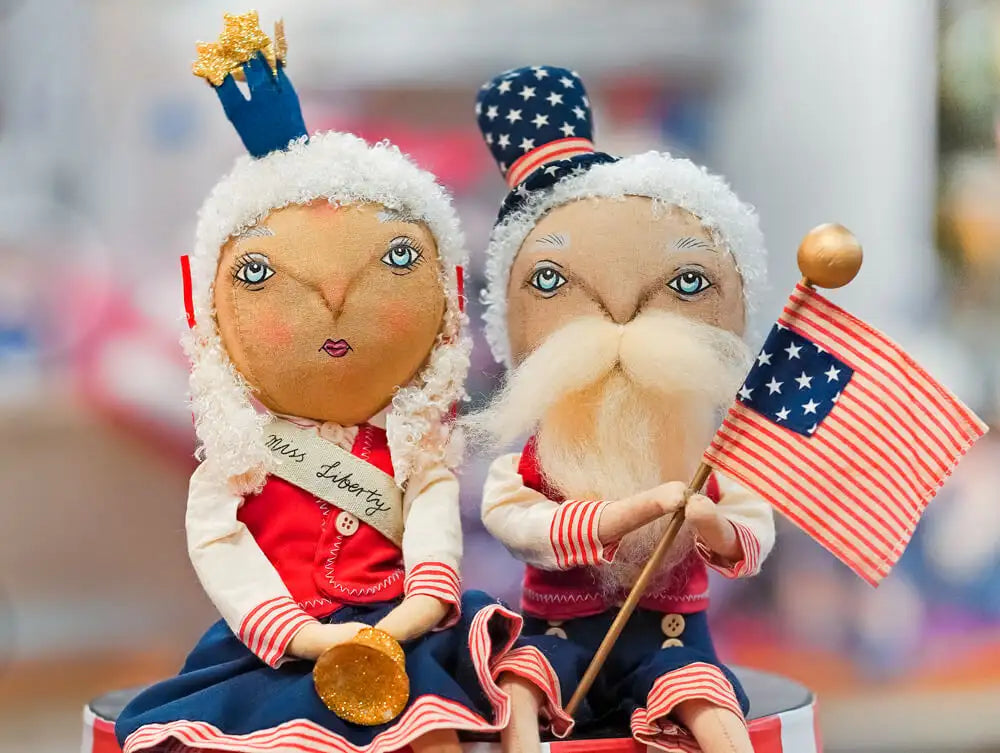 Two patriotic fabric dolls, one labeled ’miss liberty’ with a blue crown and red-and-white striped dress, the other holding an american flag with a white beard and star-spangled hat.