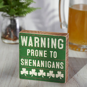 Green wooden sign with white text reading ’WARNING PRONE TO SHENANIGANS’ decorated with shamrocks.