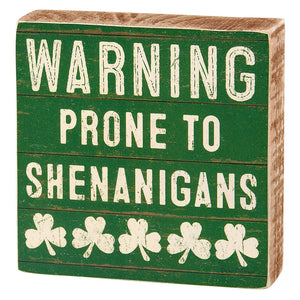 Green wooden box sign with white text reading ’WARNING PRONE TO SHENANIGANS’ and decorative shamrocks.