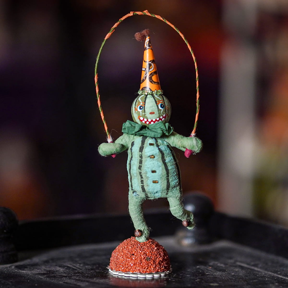 A whimsical green paper-mâché clown figurine with a spun cotton head and striped hat holding a jump rope on orange base