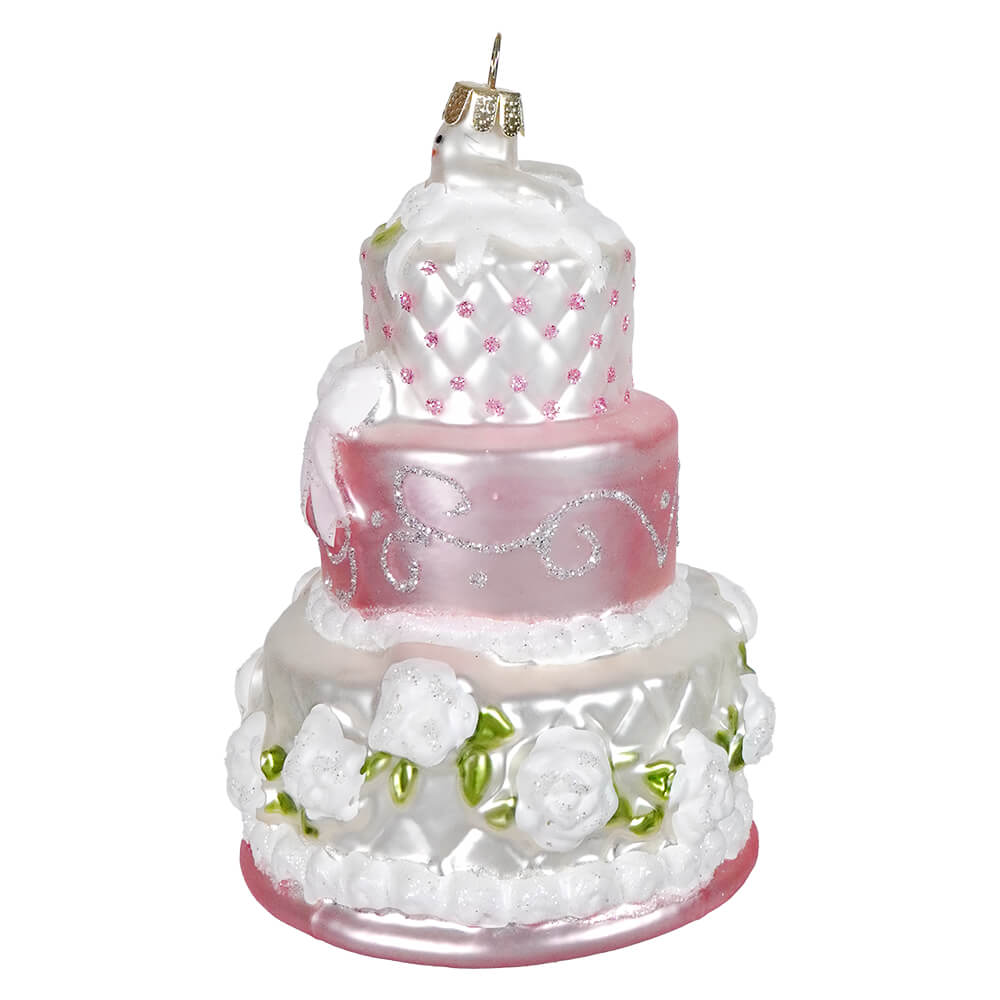 A three-tiered wedding cake ornament with white and pink layers, adorned with glittery details and floral embellishments.