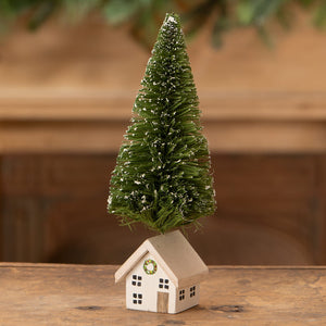 A miniature green bottle brush tree with white snow on an off-white wooden house wreath vintage christmas village trees
