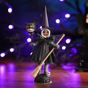 Winged spun cotton witch figure in dark green dress holding broomstick and cat mask on black base