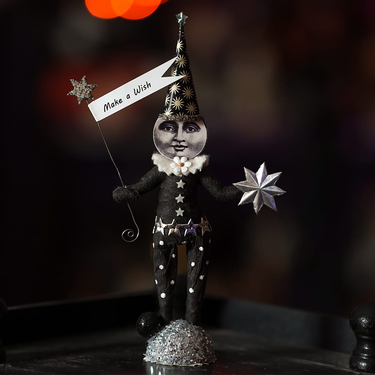 A whimsical black and silver wishing moon figure wearing a star-patterned hat and holding a make a wish banner with glittery stars