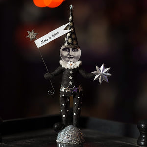 A whimsical black and silver wishing moon figure wearing a star-patterned hat and holding a make a wish banner with glittery stars