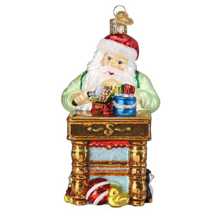 Workshop santa ornament is a glass santa ornament with red hat white beard and green coat at a gold desk with gifts