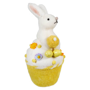 White bunny sitting atop a yellow cupcake decorated with flowers and chicks.