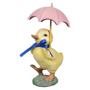 A yellow chick figurine holding pink umbrella stands on grassy base with blue ribbon around its neck